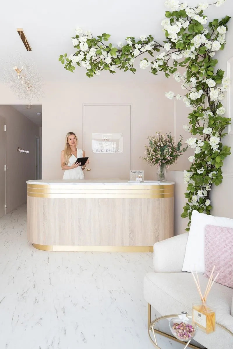 Moderne en stijlvolle receptie van een beauty- of kliniekruimte met marmeren vloer, houten balie met gouden accenten, een medewerker met tablet achter de balie en decoratieve bloesemtakken langs de muur en het plafond.