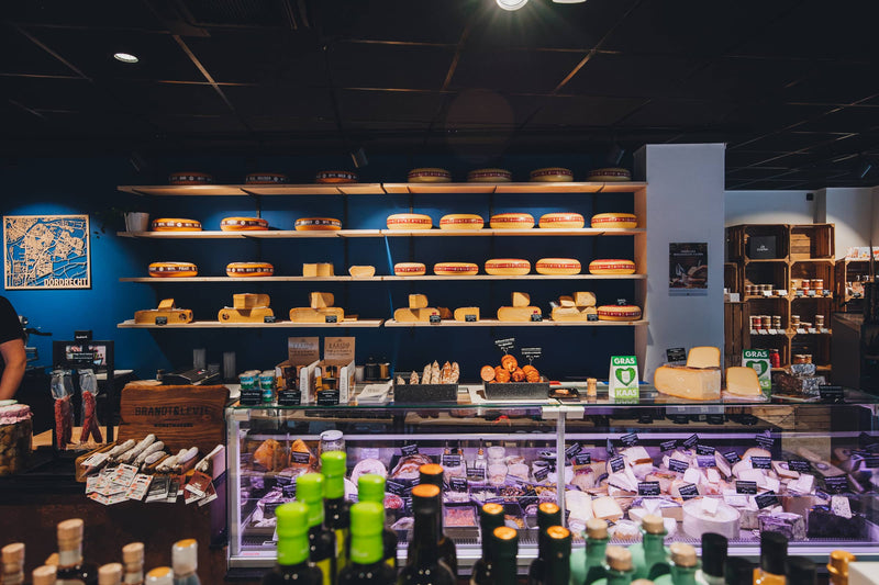 Kaaswinkel met wandpresentatie vol hele kazen, gekoelde vitrinetoonbank met diverse kazen en vleeswaren en een moderne, donkere winkelinrichting.