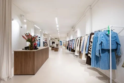 Minimalistische retail kledingwinkel met lichte inrichting, lange kledingrekken langs de muur en een houten toonbank met bloemenarrangement.