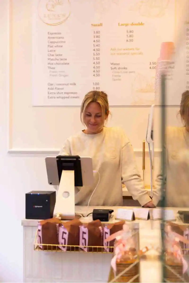 Medewerker achter de kassa in een lichte koffiezaak met menubord op de achtergrond en toonbank met verpakte producten.