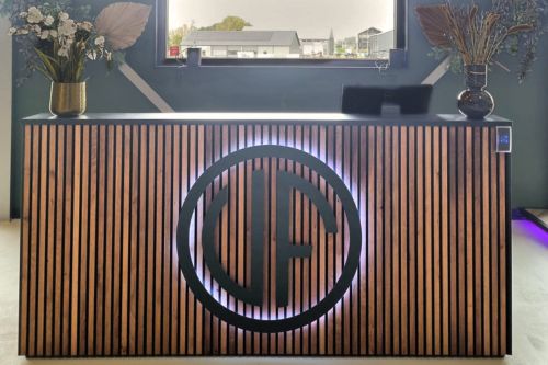 Moderne receptiebalie met houten lattenstructuur en verlicht logo aan de voorzijde, geplaatst in een stijlvolle ruimte met decoratieve vazen en een raam op de achtergrond.