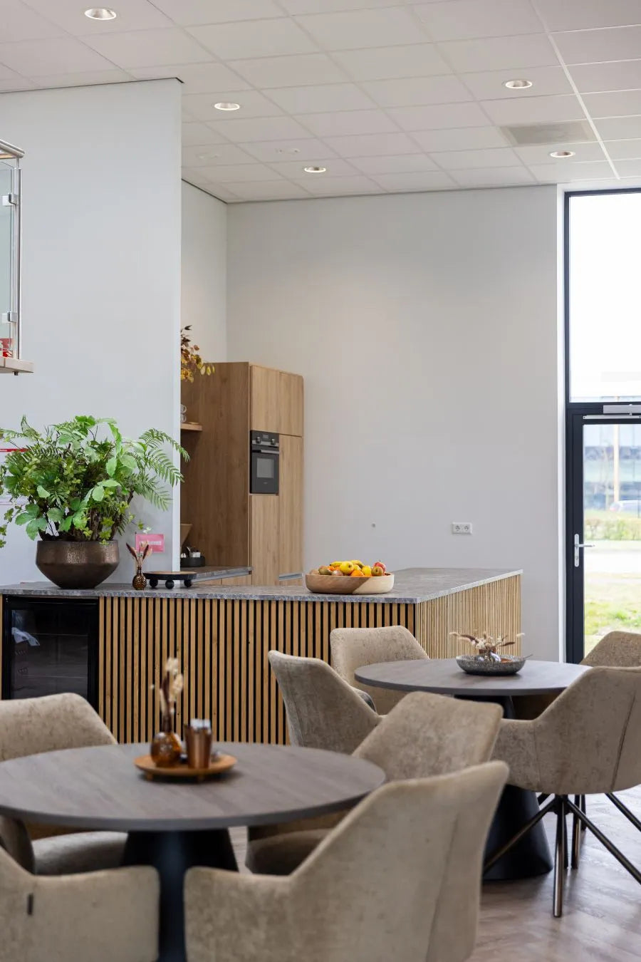 Moderne kantineruimte met ronde tafels, gestoffeerde stoelen en een houten keukenblok met lattenafwerking en decoratieve planten.