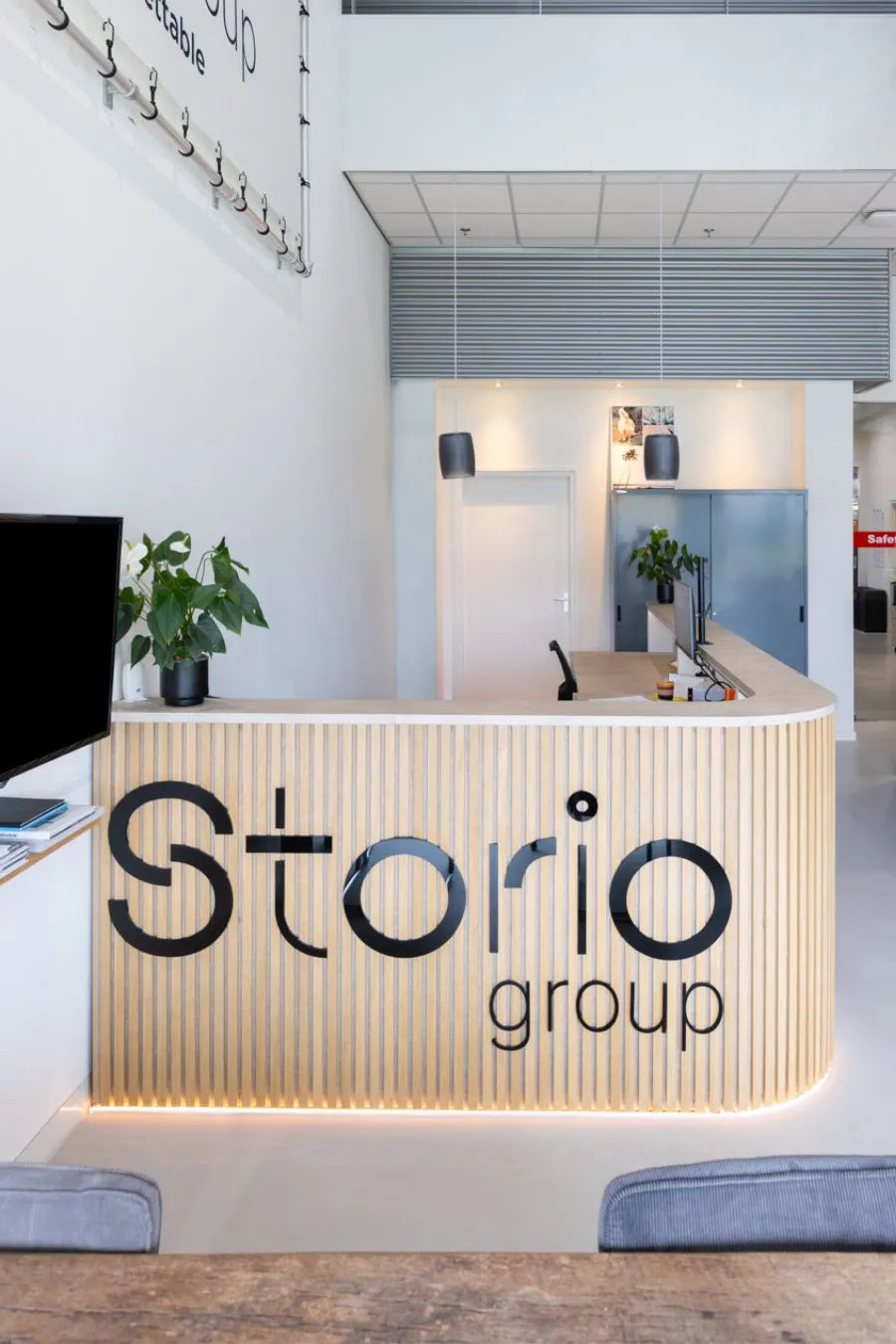 Moderne receptiebalie met houten latten en geïntegreerde verlichting, voorzien van het logo “Storio group” in een lichte, minimalistische kantoorruimte met planten en strakke afwerking.