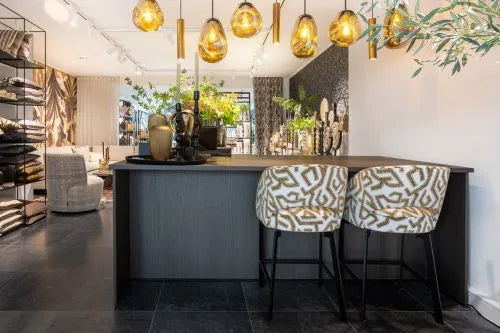 Luxe en moderne balie met donkere afwerking, stijlvolle barkrukken met patroon, warme hanglampen en decoratieve elementen, gelegen in een sfeervolle interieur- of showroomomgeving.